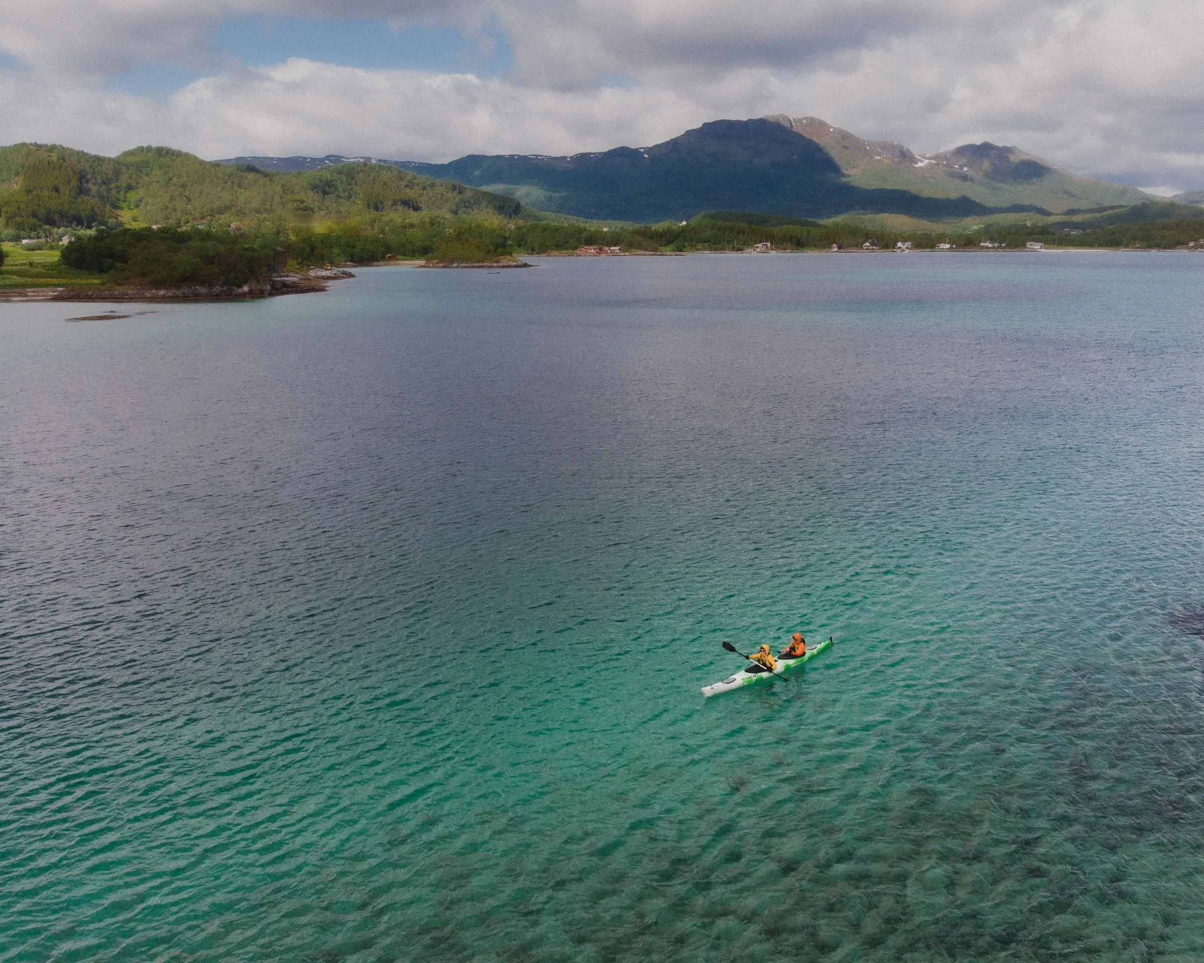 Two kayakers cross clear green water near a forested mountain shore.