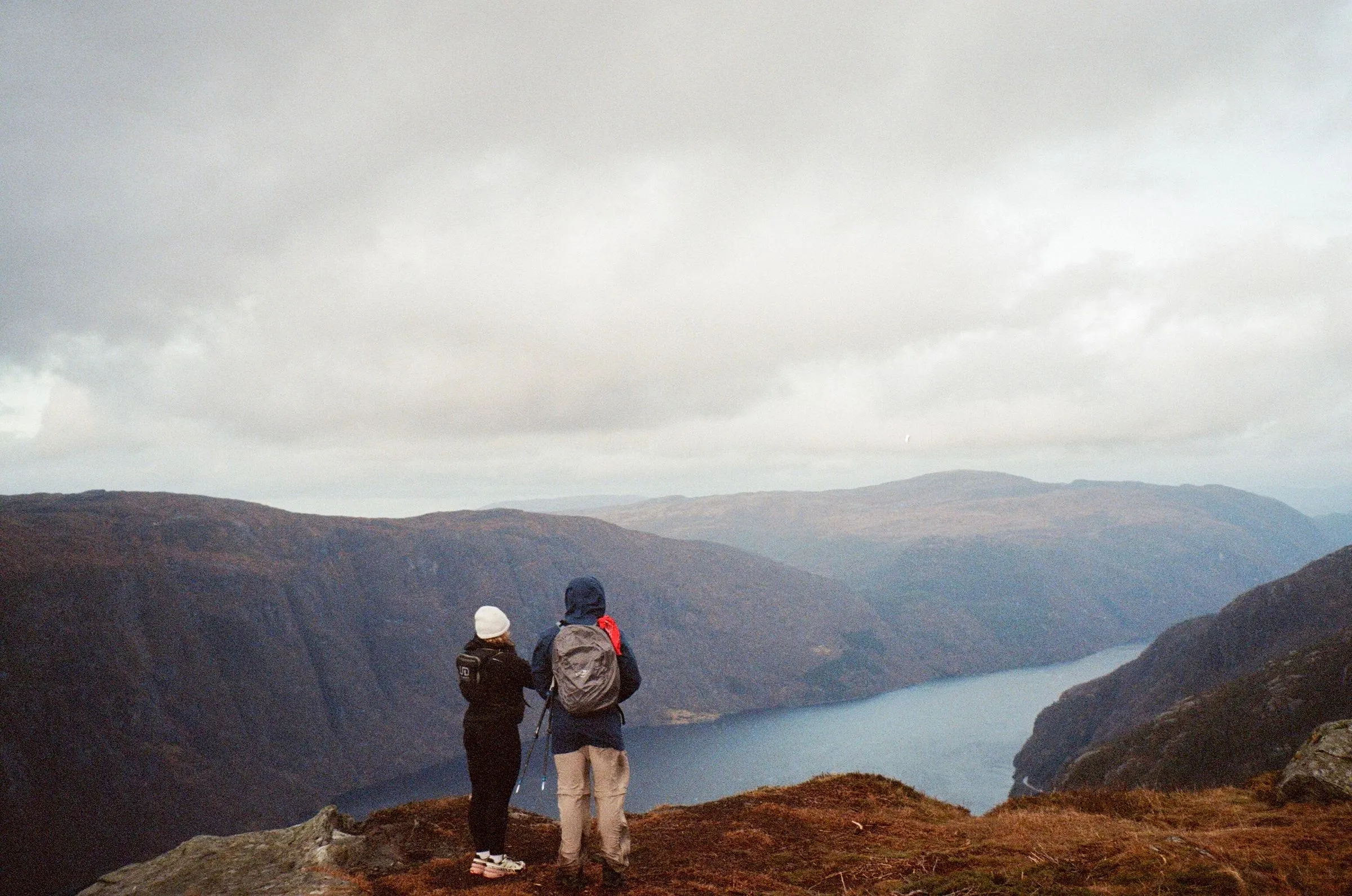 Two hikers standing on a rust-colored slope above a long blue fjord.