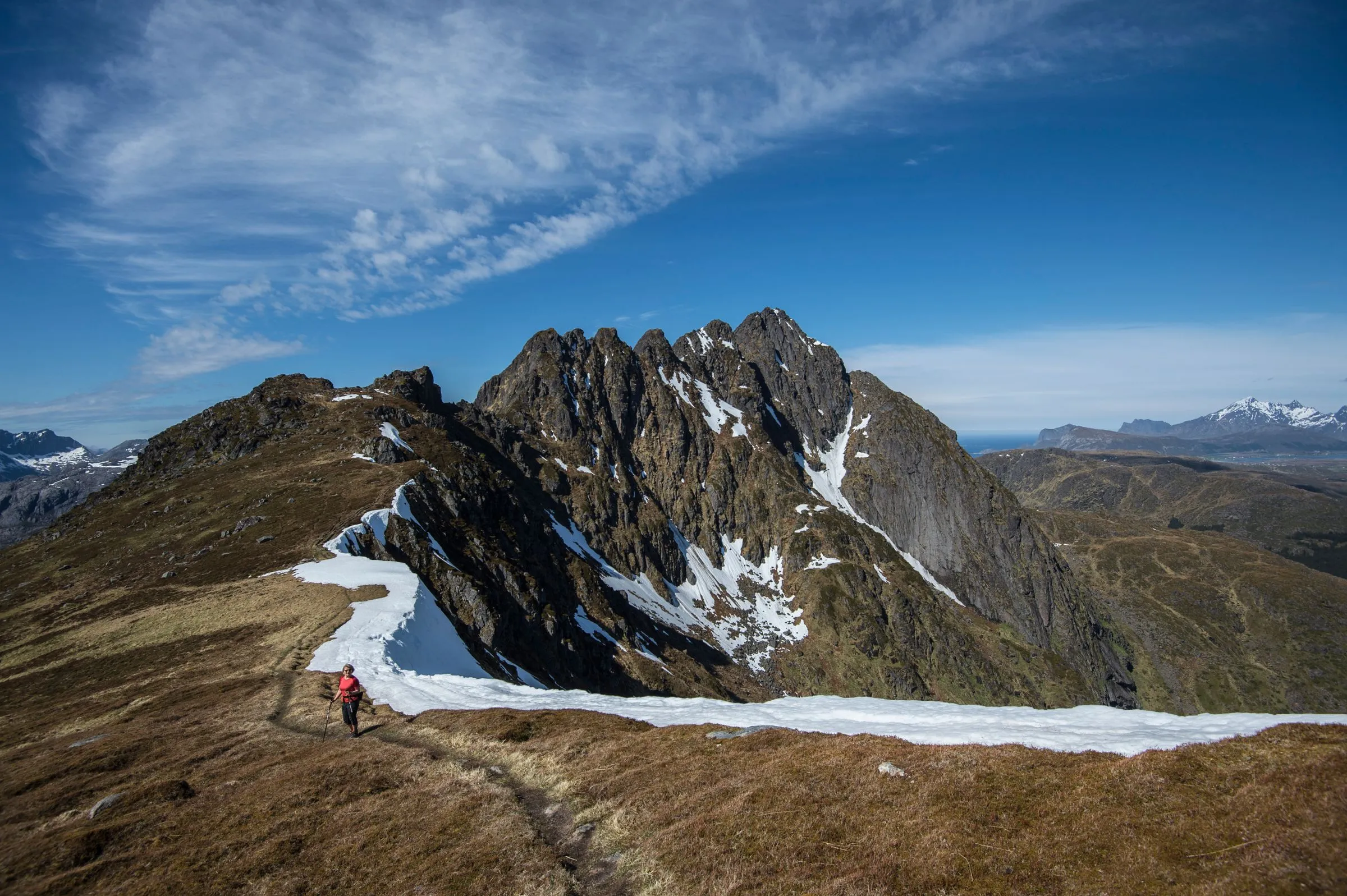 A small hiker crossing a bare mountain ridge beside patches of snow.
