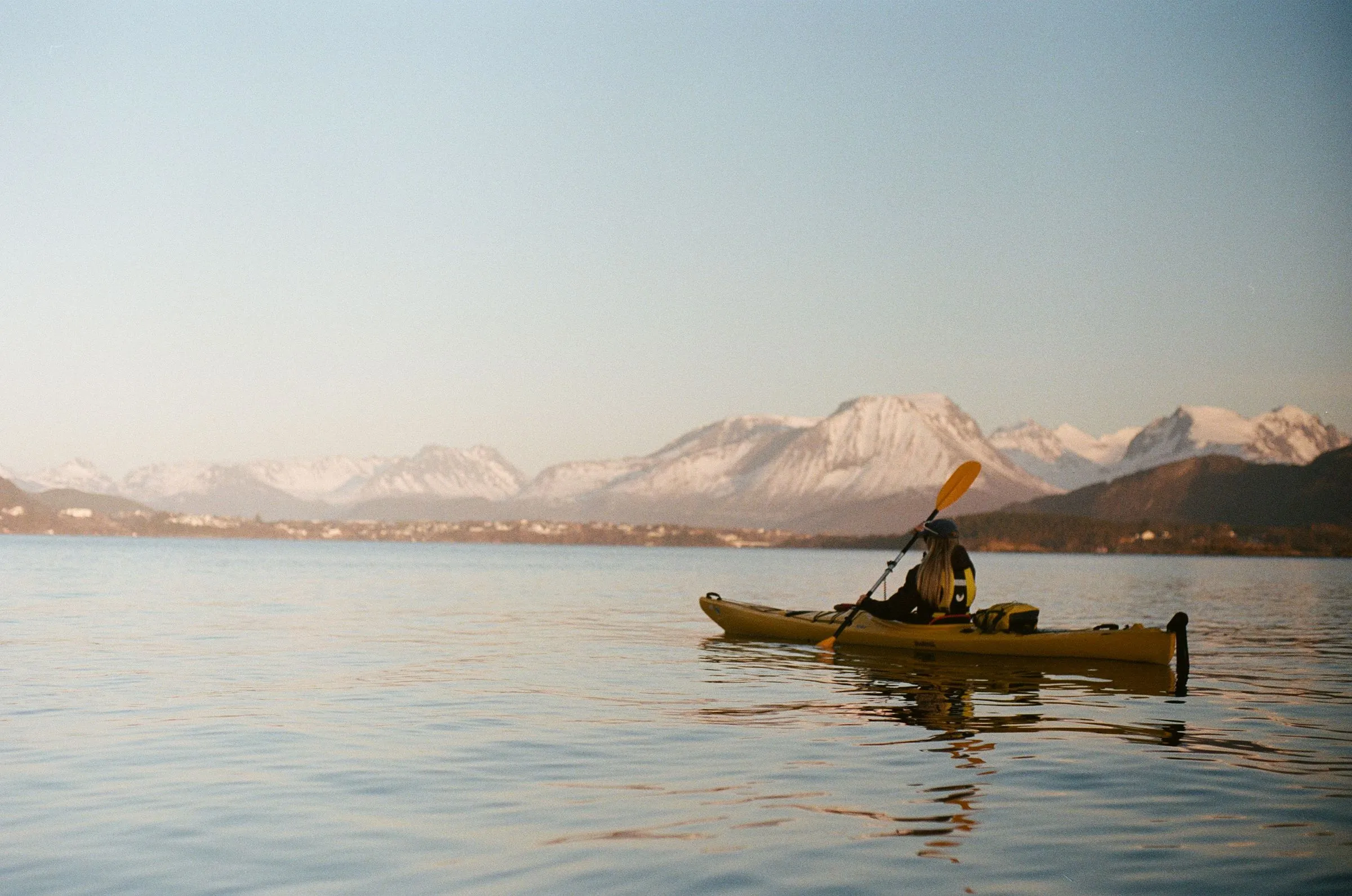 A lone sea kayaker on calm water with pale mountains across the bay.