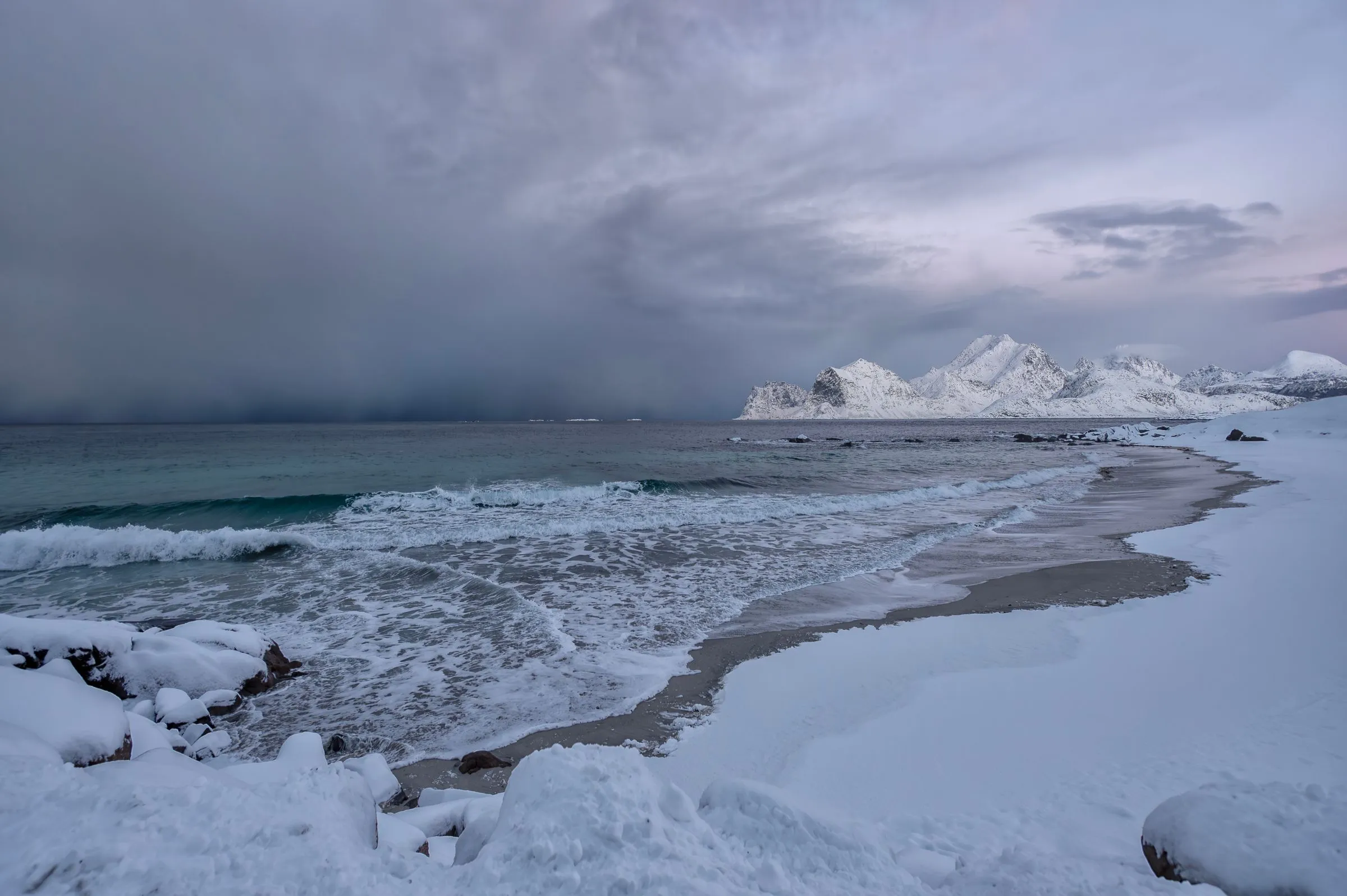 A snow-covered beach and dark winter sea beneath white coastal mountains.